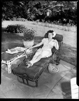 A black and white photo of Schalk outside in a bathing suit. It's a two-piece with a feathery print. The top has short sleeves and the bottom has a little skirt. She lounges on a long, padded patio chair and is smiling at the camera. In her right hand, she holds the book "Death At Her Elbow" by Donald Clough Cameron. On the table next to her are newspapers, the top of which has the headline "Big 3 Bans Nazi Race Laws." It's sunny in the background, and she wears white flowers in her hair.