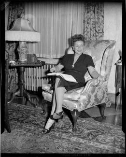 A black and white photo of Schalk, sitting in a wingback chair in a mid-century living room. She wears a knee-length, short-sleeved dark dress with a brooch at the bottom of the v-neckline. She has her legs crossed, a hardcover book open on her lap, and is smiling at something or someone off to the right.