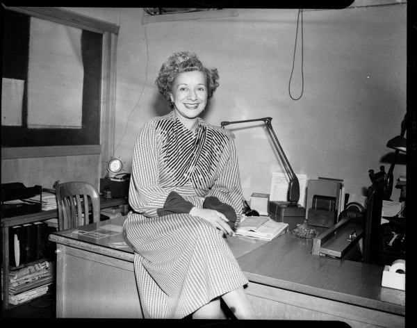 A black and white photo of Schalk, seated on her desk in the Pittsburgh Courier Newspaper office. She wears a striped, sequin dress that's long-sleeved and calf-length, and she smiles at the camera. Her hair has started to go grey and has a salt-and-pepper appearance.